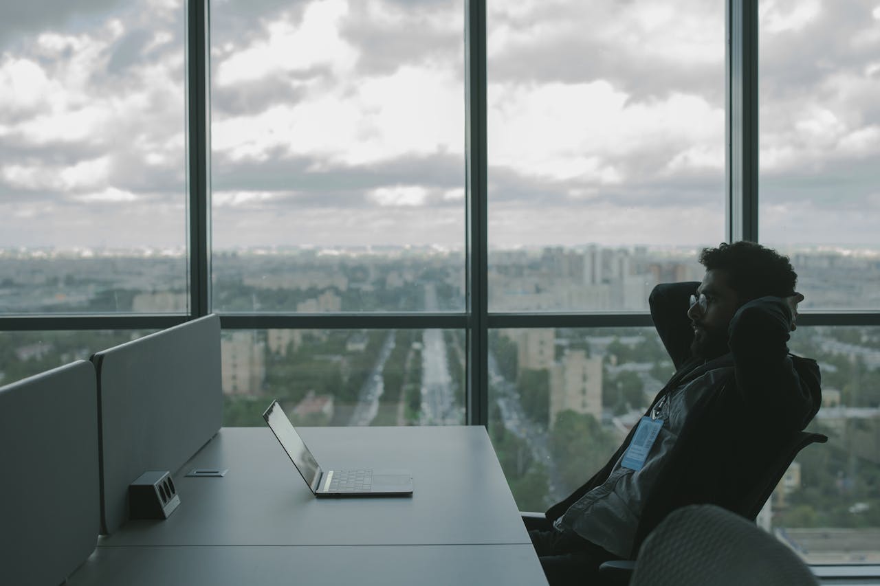 hero-img-01 A man sits in an office with hands on head in front of a laptop, overlooking a cityscape.